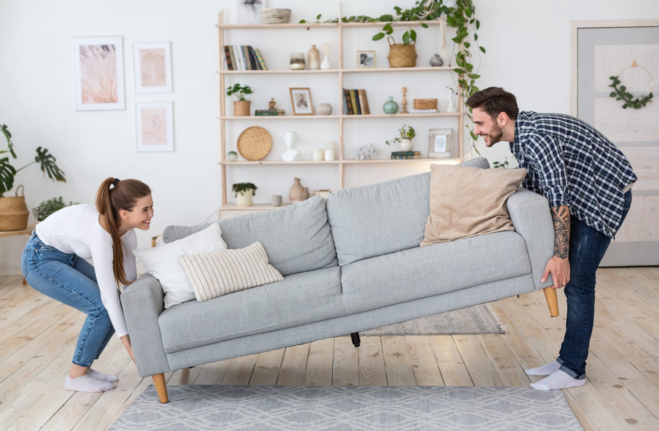 Couple lifting a sofa to prepare for Wood Floor Sanding on pale pine softwood floorboards in a bright living room.